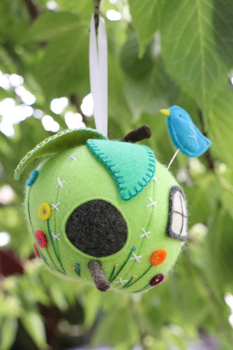 May include: A green felt apple birdhouse with a small blue bird perched on top. The apple has a round hole for the bird to enter and is decorated with embroidered flowers and white stitches.