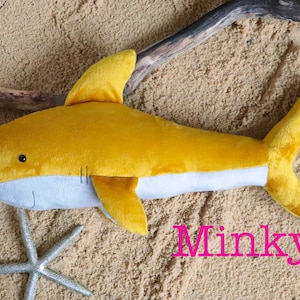 May include: A yellow plush shark toy with a gray belly, lying on a sandy surface. The toy has a small black eye and a stitched mouth. The word "Minky" is written in pink letters on the sand.