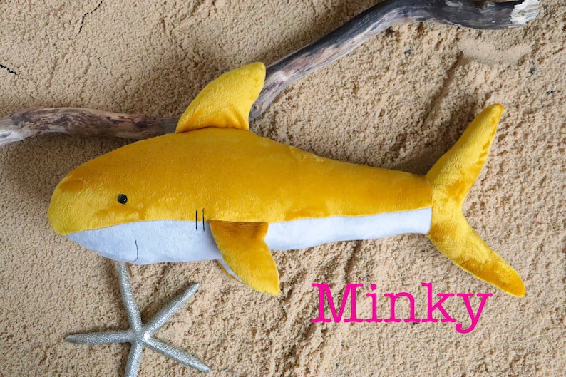 May include: A yellow plush shark toy with a gray belly, lying on a sandy surface. The toy has a small black eye and a stitched mouth. The word "Minky" is written in pink letters on the sand.