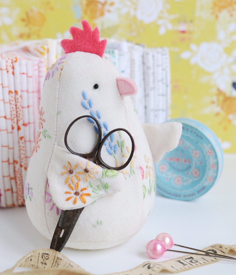 K&ouml;nnte beinhalten: Ein wei&szlig;er Stoff-H&uuml;hner-N&auml;hnadelkissen mit einem rosa Filzkamm und gestickten Blumen. Das Huhn h&auml;lt eine alte Schere und sitzt auf einer wei&szlig;en Oberfl&auml;che mit einem Ma&szlig;band und rosa N&auml;hnadeln.