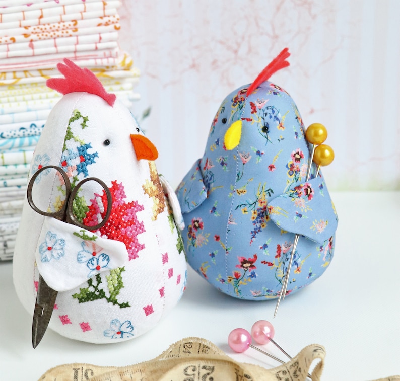 K&ouml;nnte beinhalten: Zwei Stoff-Nadelkissen in Form von H&uuml;hnern. Eines ist wei&szlig; mit roter und pinkfarbener Blumenstickerei und h&auml;lt eine Schere. Das andere ist blau mit wei&szlig;em Blumenmuster und hat goldene Stecknadeln.