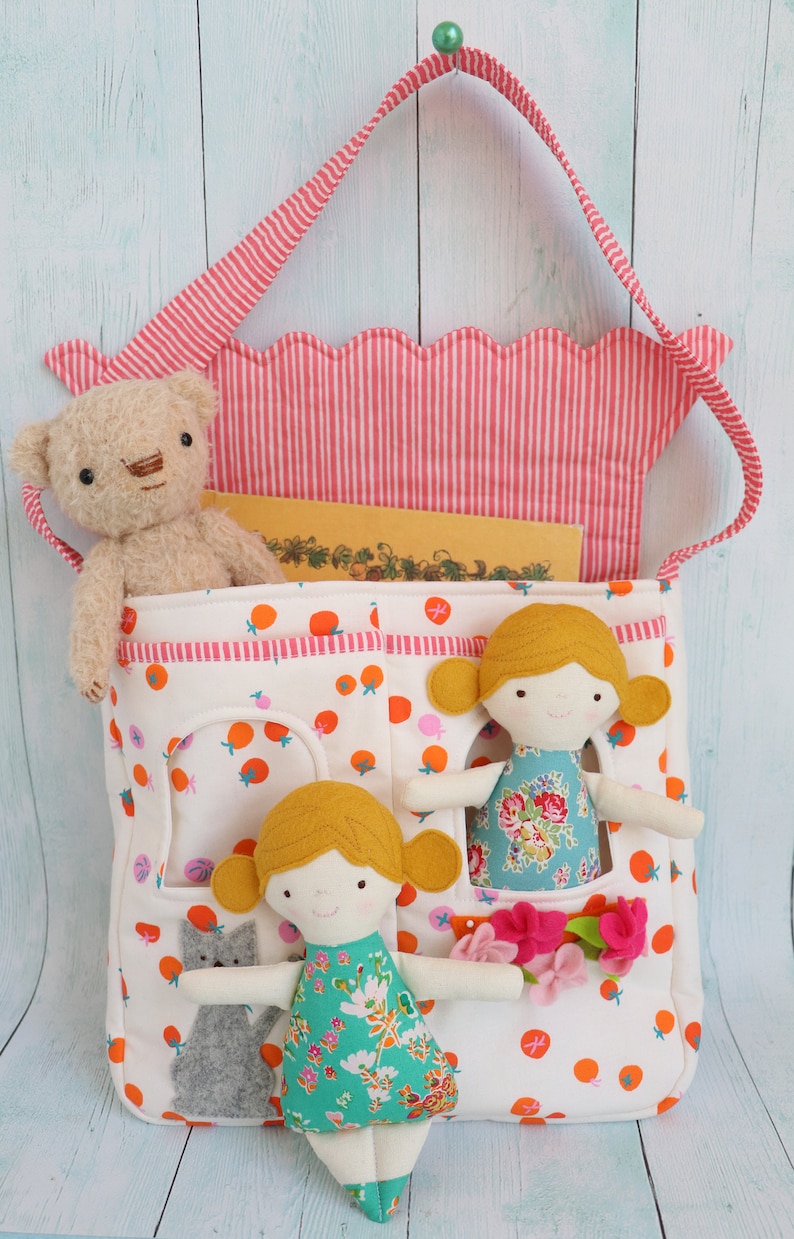 May include: A white fabric tote bag with a pink and white striped top flap. The bag has pockets on the front with two dolls and a stuffed animal inside. The dolls are wearing floral dresses and have yellow hair. The stuffed animal is a light brown teddy bear.