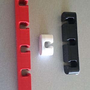 Może przedstawiać: Trzy plastikowe organizery do kabli w kolorze czerwonym, białym i czarnym. Czerwony organizer ma podłużny, prostokątny kształt z czterema szczelinami. Biały organizer to mały element w kształcie litery U. Czarny organizer jest podobny do czerwonego.