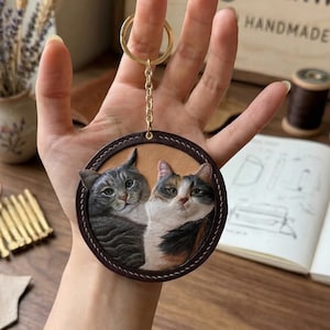 May include: A round leather keychain featuring a detailed portrait of two cats. The cats are gray tabby and calico, set against a tan background with a dark brown border. The keychain has a gold-colored chain and ring.