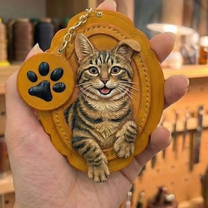 Puede incluir: Un llavero de cuero marr&oacute;n con un dise&ntilde;o de gato atigrado en relieve 3D. El gato est&aacute; sonriendo y enmarcado dentro de un &oacute;valo. Un peque&ntilde;o dije redondo con una huella de pata negra cuelga de una cadena dorada.