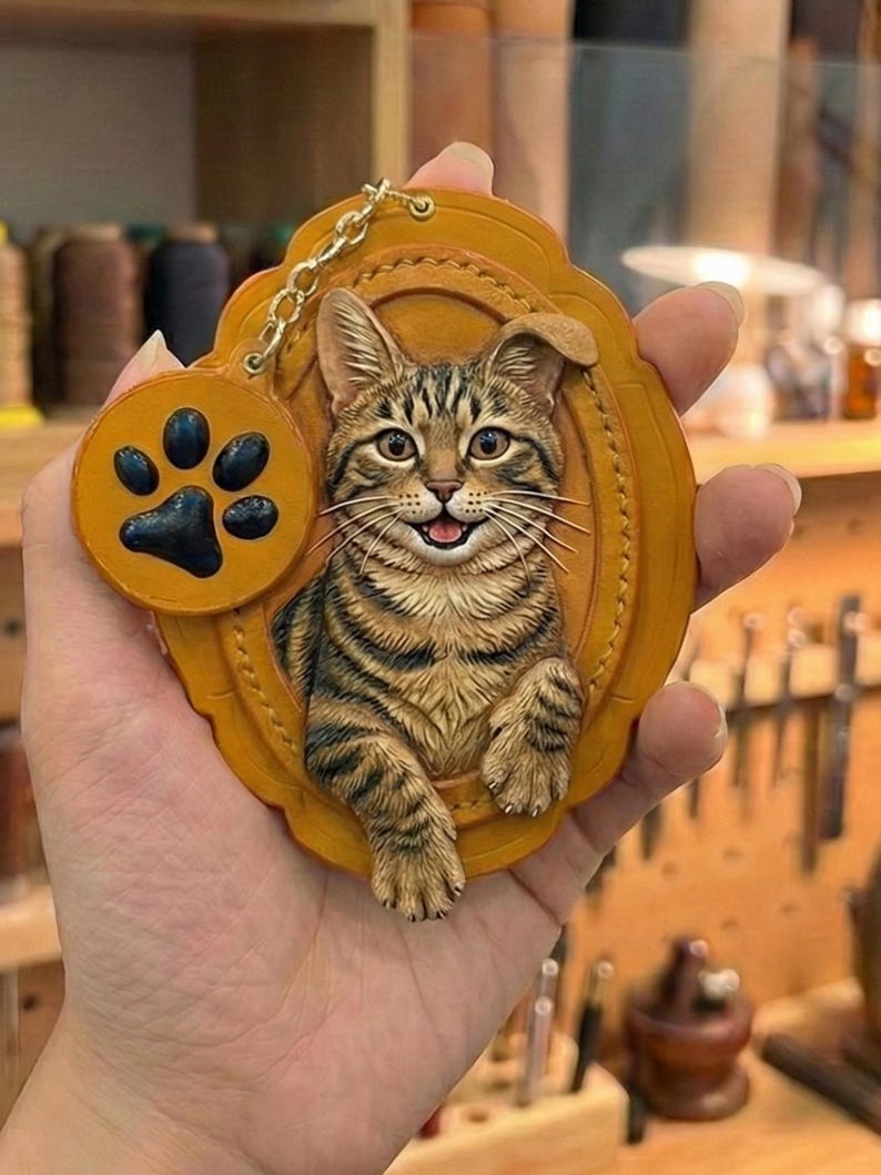Puede incluir: Un llavero de cuero marr&oacute;n con un dise&ntilde;o de gato atigrado en relieve 3D. El gato est&aacute; sonriendo y enmarcado dentro de un &oacute;valo. Un peque&ntilde;o dije redondo con una huella de pata negra cuelga de una cadena dorada.