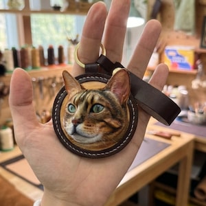 Puede incluir: Un llavero redondo de cuero con un retrato detallado de la cara de un gato con ojos verdes y pelaje marr&oacute;n y beige. El llavero tiene una correa de cuero marr&oacute;n y un anillo de metal dorado.