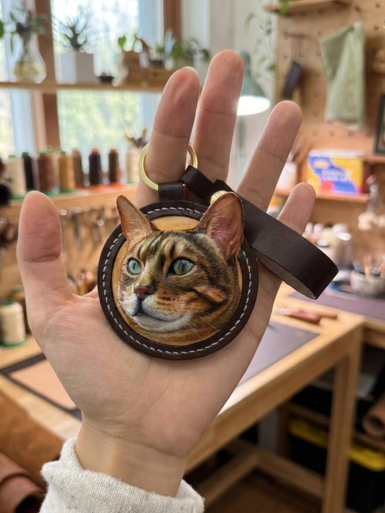 Puede incluir: Un llavero redondo de cuero con un retrato detallado de la cara de un gato con ojos verdes y pelaje marr&oacute;n y beige. El llavero tiene una correa de cuero marr&oacute;n y un anillo de metal dorado.