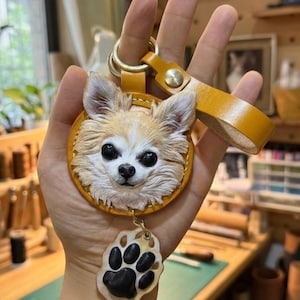 Puede incluir: Llavero de cuero hecho a mano con un detallado rostro de perro chihuahua en tonos beige y blanco. El llavero tiene un anillo de color dorado y una correa de cuero a juego. Un dije de huella de pata cuelga debajo de la cara del perro.