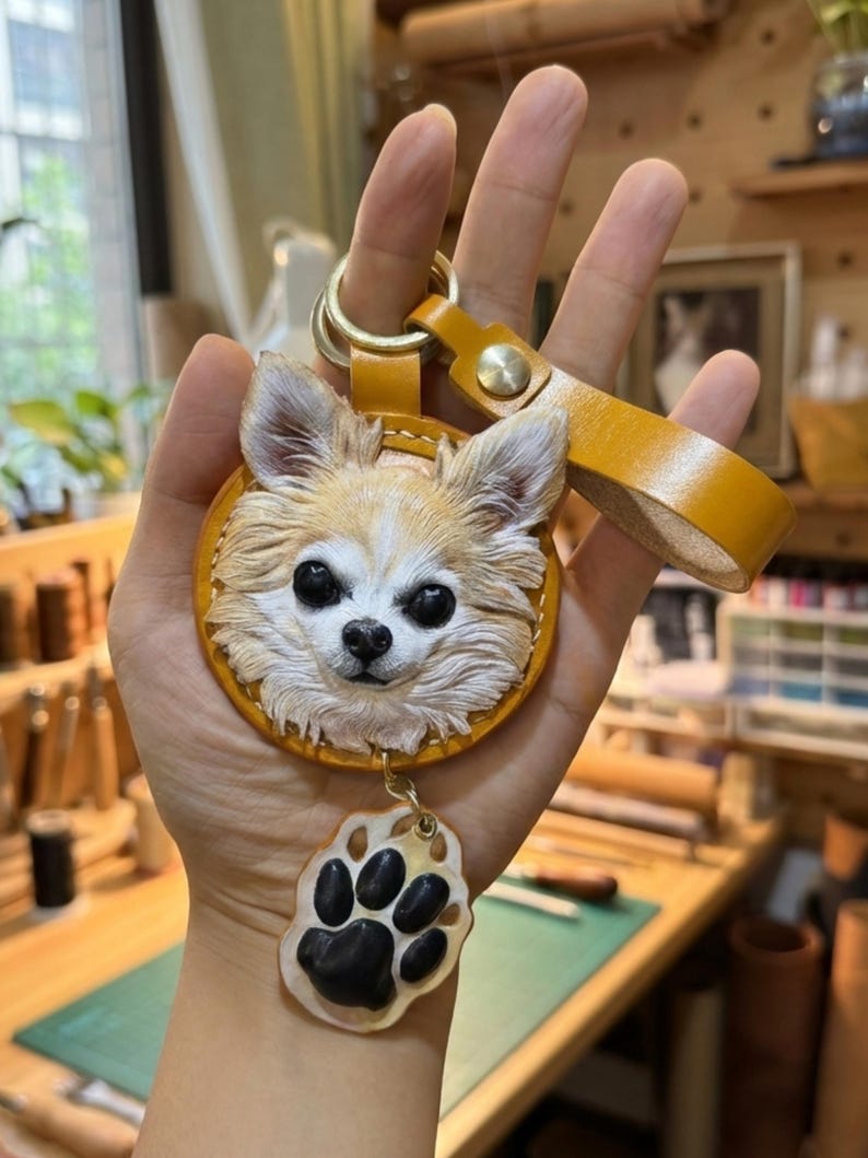 Puede incluir: Llavero de cuero hecho a mano con un detallado rostro de perro chihuahua en tonos beige y blanco. El llavero tiene un anillo de color dorado y una correa de cuero a juego. Un dije de huella de pata cuelga debajo de la cara del perro.
