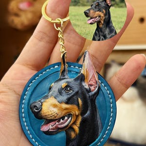 Puede incluir: Un llavero de Doberman Pinscher con un dise&ntilde;o de cuero repujado detallado. La cabeza del perro es negra y marr&oacute;n, sobre un fondo circular azul con costuras blancas. Una cadena y un anillo dorados completan el accesorio.