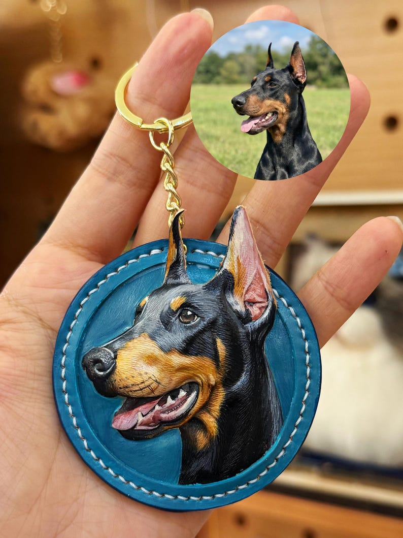 Puede incluir: Un llavero de Doberman Pinscher con un dise&ntilde;o de cuero repujado detallado. La cabeza del perro es negra y marr&oacute;n, sobre un fondo circular azul con costuras blancas. Una cadena y un anillo dorados completan el accesorio.