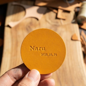 Puede incluir: Un posavasos redondo de cuero amarillo mostaza con el nombre "Naru" y la fecha "2025.10.24" grabados en relieve. El posavasos se sostiene en una mano, con una superficie de madera y herramientas de cuero en el fondo.