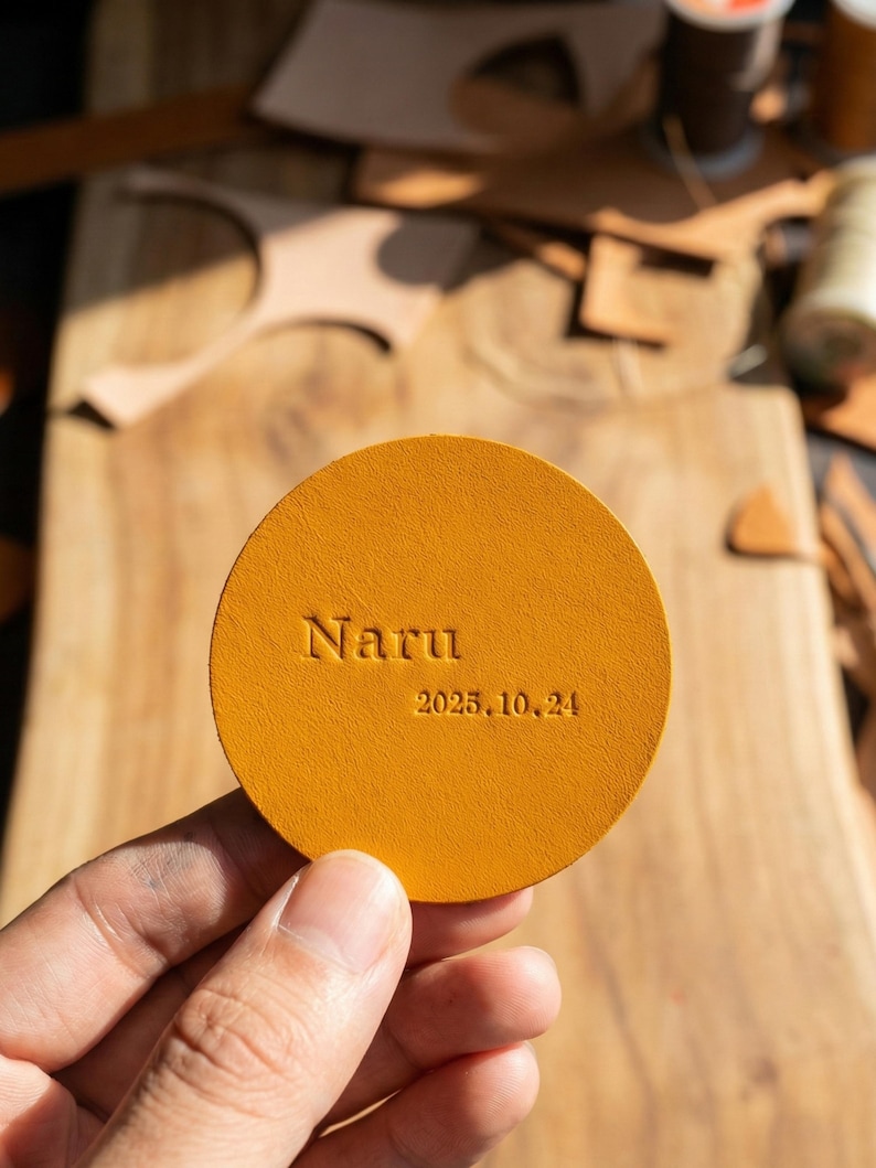 Puede incluir: Un posavasos redondo de cuero amarillo mostaza con el nombre "Naru" y la fecha "2025.10.24" grabados en relieve. El posavasos se sostiene en una mano, con una superficie de madera y herramientas de cuero en el fondo.