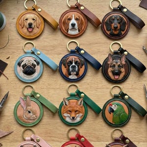 Puede incluir: Colecci&oacute;n de llaveros de cuero hechos a mano con retratos detallados de animales. Cada llavero tiene un dise&ntilde;o circular con un anillo dorado y una correa de cuero a juego. Animales: perros, un conejo, un zorro, un loro, un h&aacute;mster, una ardilla y un b&uacute;ho.