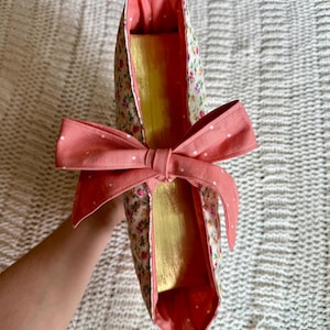 K&ouml;nnte beinhalten: Eine Tragetasche mit floralem Muster und einer korallenfarbenen Schleife. Die Tasche hat ein goldenes Innenfutter und ein korallenfarbenes Futter. Der Au&szlig;enstoff hat ein Muster aus kleinen Blumen und Bl&auml;ttern auf cremefarbenem Hintergrund. Die Schleife ist mit Wei&szlig; gepunktet.
