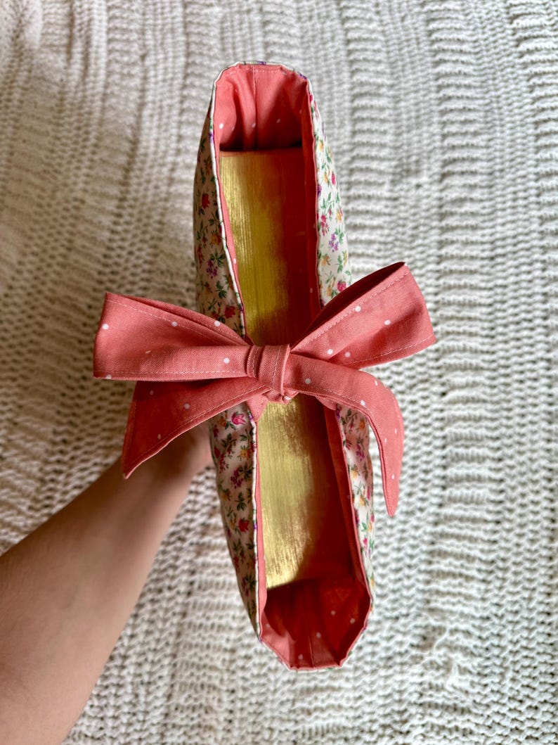 K&ouml;nnte beinhalten: Eine Tragetasche mit floralem Muster und einer korallenfarbenen Schleife. Die Tasche hat ein goldenes Innenfutter und ein korallenfarbenes Futter. Der Au&szlig;enstoff hat ein Muster aus kleinen Blumen und Bl&auml;ttern auf cremefarbenem Hintergrund. Die Schleife ist mit Wei&szlig; gepunktet.