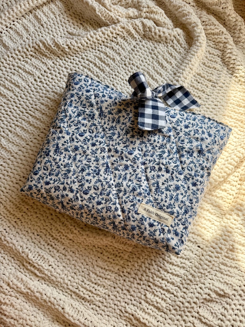 K&ouml;nnte beinhalten: Quadratisches, gestepptes Kissen mit blauem Blumenmuster auf wei&szlig;em Grund. Oben ist eine kleine Schleife aus blau-wei&szlig; kariertem Stoff befestigt. Das Etikett lautet "A Faith Original".