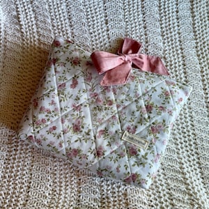 K&ouml;nnte beinhalten: Ein gestepptes Kissen mit floralem Muster und einer rosa Schleife. Das Kissen hat einen wei&szlig;en Hintergrund mit einem Muster aus rosa Rosen und gr&uuml;nen Bl&auml;ttern. Die Schleife ist einfarbig rosa. Der Text "A Faith Original" befindet sich auf einem kleinen Etikett.