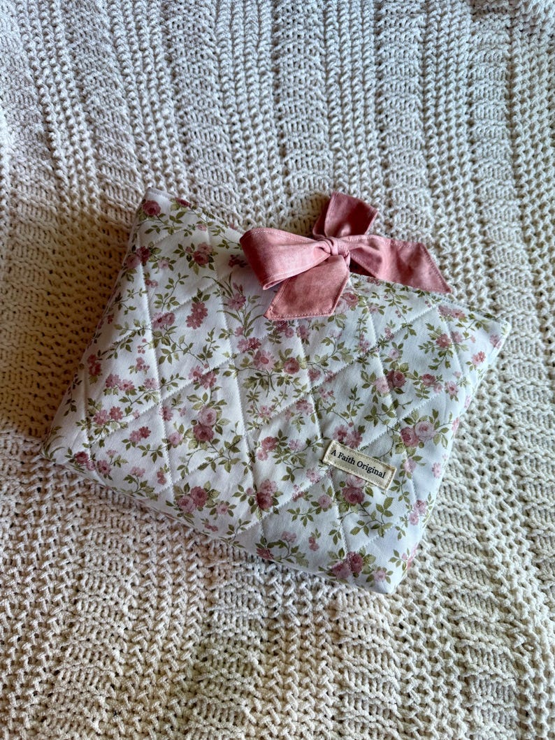 K&ouml;nnte beinhalten: Ein gestepptes Kissen mit floralem Muster und einer rosa Schleife. Das Kissen hat einen wei&szlig;en Hintergrund mit einem Muster aus rosa Rosen und gr&uuml;nen Bl&auml;ttern. Die Schleife ist einfarbig rosa. Der Text "A Faith Original" befindet sich auf einem kleinen Etikett.