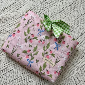 K&ouml;nnte beinhalten: Eine gesteppte rosa Tasche mit Erdbeer- und Blumenmuster, blauen Schleifen und einer gr&uuml;n-wei&szlig; karierten Schleife. Die Tasche tr&auml;gt das Etikett "A Faith Original".