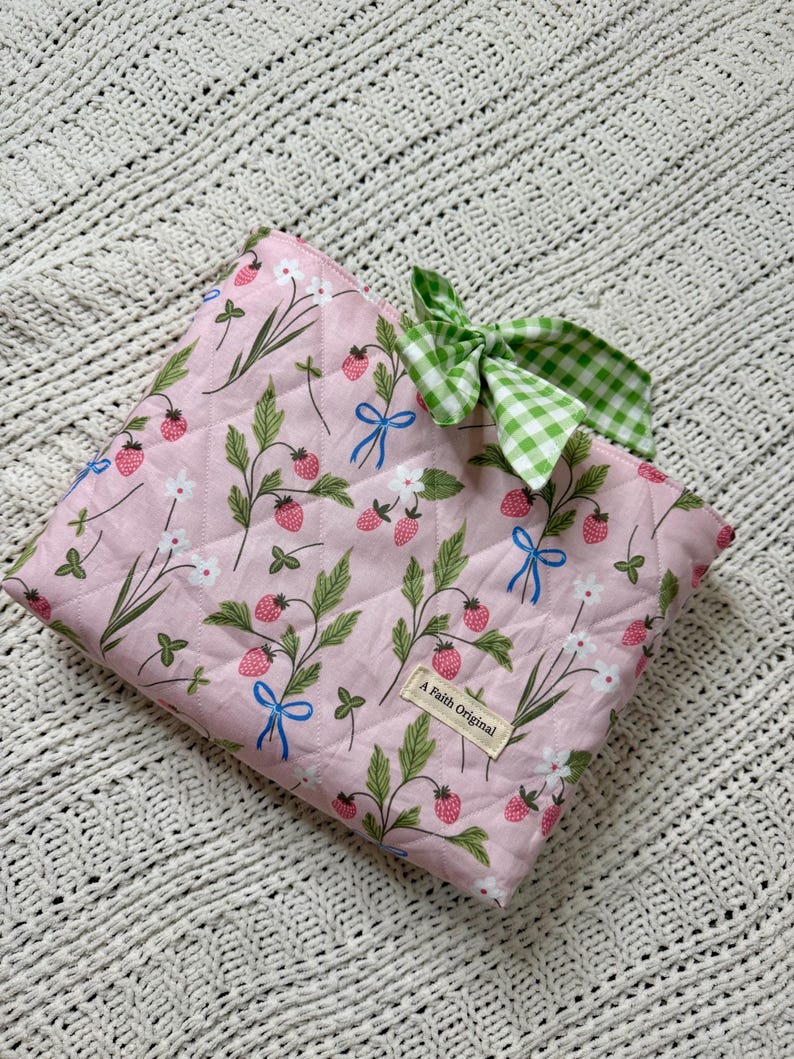 K&ouml;nnte beinhalten: Eine gesteppte rosa Tasche mit Erdbeer- und Blumenmuster, blauen Schleifen und einer gr&uuml;n-wei&szlig; karierten Schleife. Die Tasche tr&auml;gt das Etikett "A Faith Original".