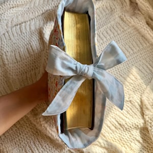 K&ouml;nnte beinhalten: Eine Buchh&uuml;lle aus floralem Stoff mit einer hellblauen Vichy-Schleife. Die H&uuml;lle weist ein Muster aus kleinen Blumen in Rosa-, Orange- und Gelbt&ouml;nen auf. Die Seiten des Buches haben einen goldfarbenen Rand.