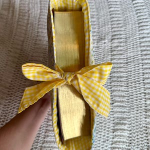 K&ouml;nnte beinhalten: Eine Bucheinbandtasche mit gelber Vichy-Schleife. Die Tasche ist mit gelbem Vichy-Stoff und einem Blumenmuster gef&uuml;ttert. Die Seiten des Buches haben einen goldenen Farbton.