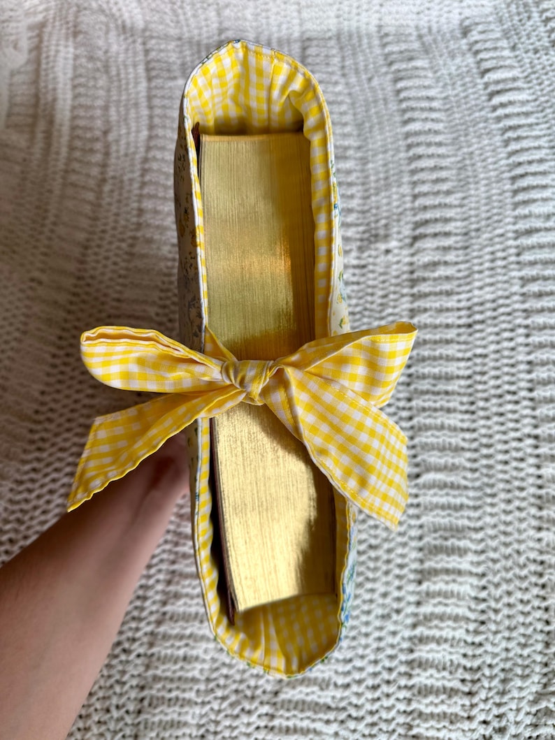K&ouml;nnte beinhalten: Eine Bucheinbandtasche mit gelber Vichy-Schleife. Die Tasche ist mit gelbem Vichy-Stoff und einem Blumenmuster gef&uuml;ttert. Die Seiten des Buches haben einen goldenen Farbton.