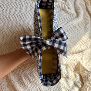 K&ouml;nnte beinhalten: Ein handgefertigter Schuh mit blauem Blumenmuster und einer Vichy-Schleife. Die Innenseite ist goldfarben. Der Schuh wird vor einem cremefarbenen, strukturierten Hintergrund pr&auml;sentiert.
