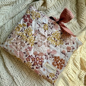 K&ouml;nnte beinhalten: Eine gesteppte Kissenh&uuml;lle mit floralem Muster und Schleife. Die H&uuml;lle hat einen wei&szlig;en Hintergrund mit einem Muster aus rosa, braunen und goldenen Blumen. Die Schleife ist einfarbig rosa mit wei&szlig;en Punkten. Das Etikett lautet "A Faith Original."