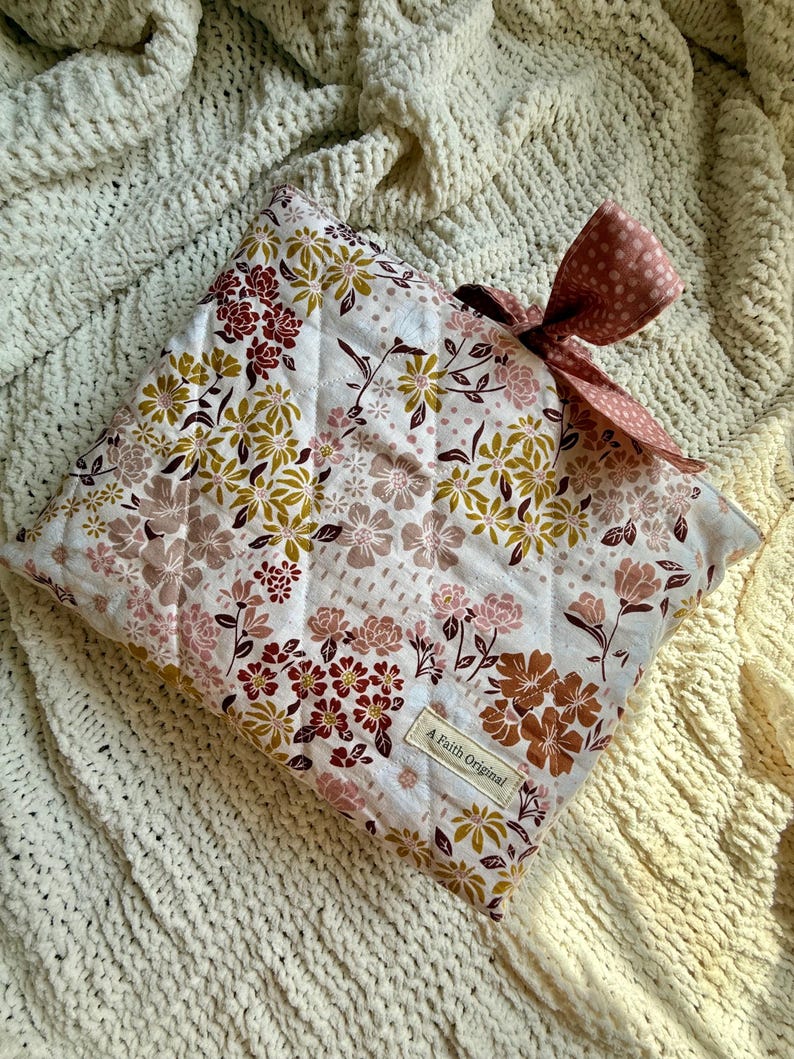 K&ouml;nnte beinhalten: Eine gesteppte Kissenh&uuml;lle mit floralem Muster und Schleife. Die H&uuml;lle hat einen wei&szlig;en Hintergrund mit einem Muster aus rosa, braunen und goldenen Blumen. Die Schleife ist einfarbig rosa mit wei&szlig;en Punkten. Das Etikett lautet "A Faith Original."