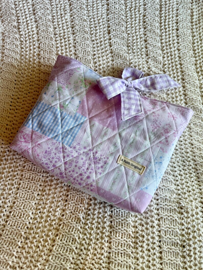 K&ouml;nnte beinhalten: Gesteppte Patchwork-Tasche in Lavendel-, Rosa- und Blaut&ouml;nen mit einer Vichy-Schleife. Die Tasche hat ein Rautenmuster und ein Etikett mit der Aufschrift "A Faith Original".