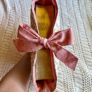 K&ouml;nnte beinhalten: Eine Buchh&uuml;lle aus floralem Stoff mit einer rosa Schleife. Die H&uuml;lle ist wei&szlig; mit einem zarten Blumenmuster und einem rosa Innenfutter. Die Schleife ist einfarbig rosa. Die goldfarbenen Seiten des Buches sind sichtbar.