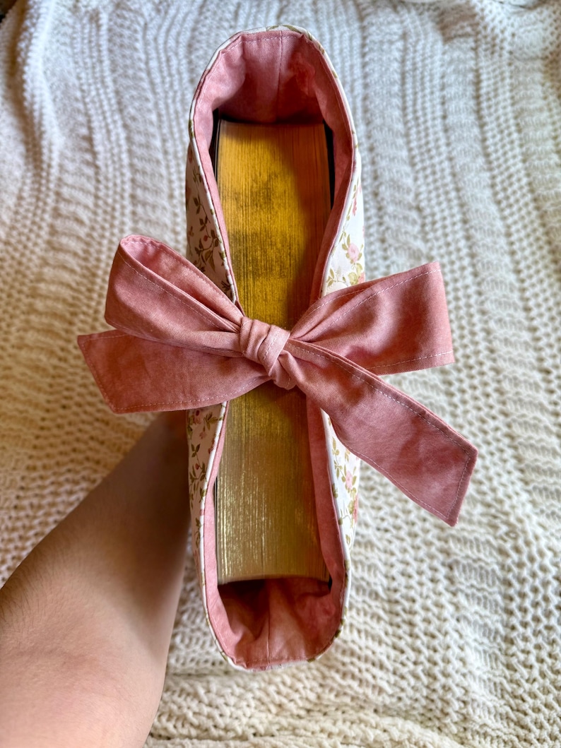 K&ouml;nnte beinhalten: Eine Buchh&uuml;lle aus floralem Stoff mit einer rosa Schleife. Die H&uuml;lle ist wei&szlig; mit einem zarten Blumenmuster und einem rosa Innenfutter. Die Schleife ist einfarbig rosa. Die goldfarbenen Seiten des Buches sind sichtbar.