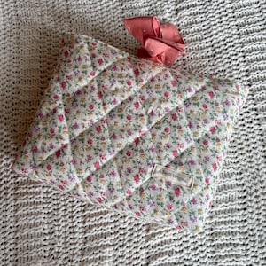 K&ouml;nnte beinhalten: Gesteppte H&uuml;lle mit floralem Druck und einer rosa Schleife. Die H&uuml;lle hat einen wei&szlig;en Hintergrund mit kleinen rosa, lila und gelben Blumen. Das Etikett lautet "A Faith Original".