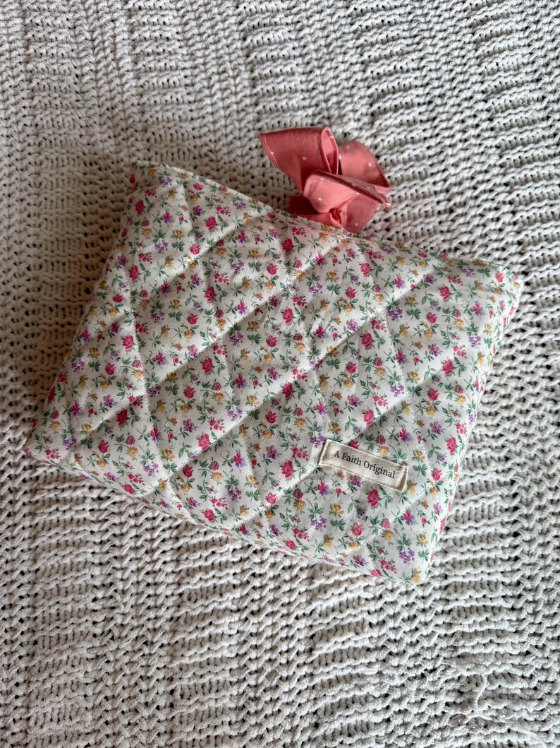 K&ouml;nnte beinhalten: Gesteppte H&uuml;lle mit floralem Druck und einer rosa Schleife. Die H&uuml;lle hat einen wei&szlig;en Hintergrund mit kleinen rosa, lila und gelben Blumen. Das Etikett lautet "A Faith Original".