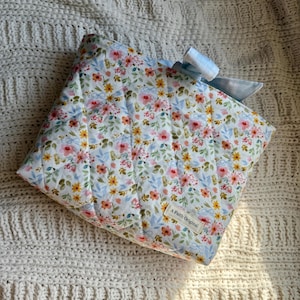 K&ouml;nnte beinhalten: Eine gesteppte Tasche mit floralem Muster und einer hellblauen Vichy-Schleife. Die Tasche hat einen wei&szlig;en Hintergrund mit rosa, gelben und blauen Blumen. Das Etikett lautet "A Faith Original".