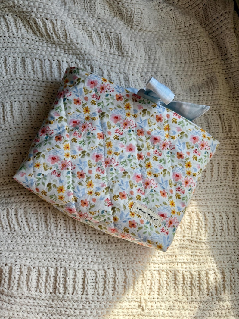 K&ouml;nnte beinhalten: Eine gesteppte Tasche mit floralem Muster und einer hellblauen Vichy-Schleife. Die Tasche hat einen wei&szlig;en Hintergrund mit rosa, gelben und blauen Blumen. Das Etikett lautet "A Faith Original".