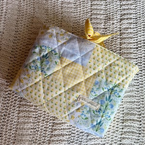 K&ouml;nnte beinhalten: Gesteppte Stofftasche mit Patchwork-Design in gelben, blauen und wei&szlig;en Blumenmustern. Oben ist ein gelbes Vichy-Band gebunden. Das Etikett lautet "A Faith Original".