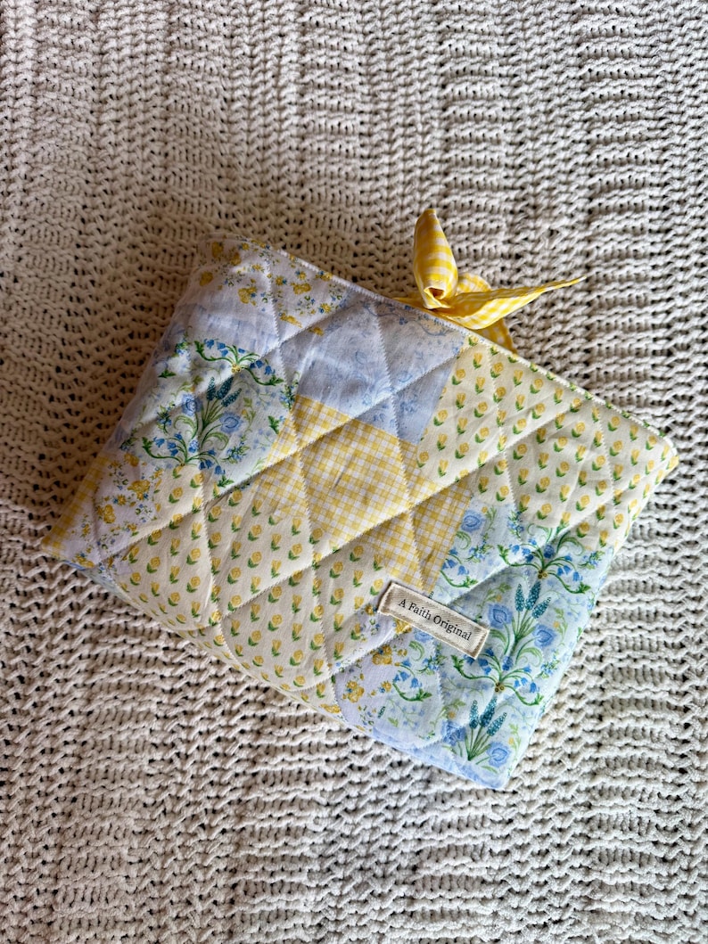 K&ouml;nnte beinhalten: Gesteppte Stofftasche mit Patchwork-Design in gelben, blauen und wei&szlig;en Blumenmustern. Oben ist ein gelbes Vichy-Band gebunden. Das Etikett lautet "A Faith Original".