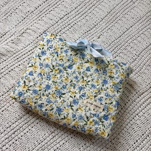 K&ouml;nnte beinhalten: Eine quadratische Tasche mit floralem Muster aus blauen und gelben Blumen auf wei&szlig;em Grund. Sie hat eine hellblaue Schleife und ein Etikett mit dem Text "A Faith Original". Die Tasche liegt auf einer wei&szlig;en, strukturierten Decke.