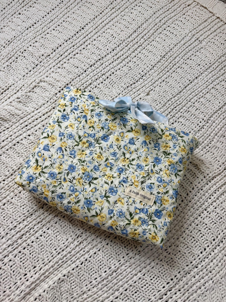K&ouml;nnte beinhalten: Eine quadratische Tasche mit floralem Muster aus blauen und gelben Blumen auf wei&szlig;em Grund. Sie hat eine hellblaue Schleife und ein Etikett mit dem Text "A Faith Original". Die Tasche liegt auf einer wei&szlig;en, strukturierten Decke.