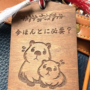 以下が含まれることがあります： バッグにぶら下がっている、クマのイラストと日本語のテキストが入った木製のタグ。タグは茶色で、2匹のクマと日本語のテキストが彫られています。バッグは青と茶色です。