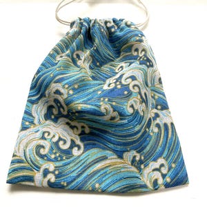 以下が含まれることがあります： 青と白の波模様の巾着袋。生地は、青、白、金色の色合いで様式化された海の波の繰り返しデザインが特徴です。袋はベージュの巾着留めです。