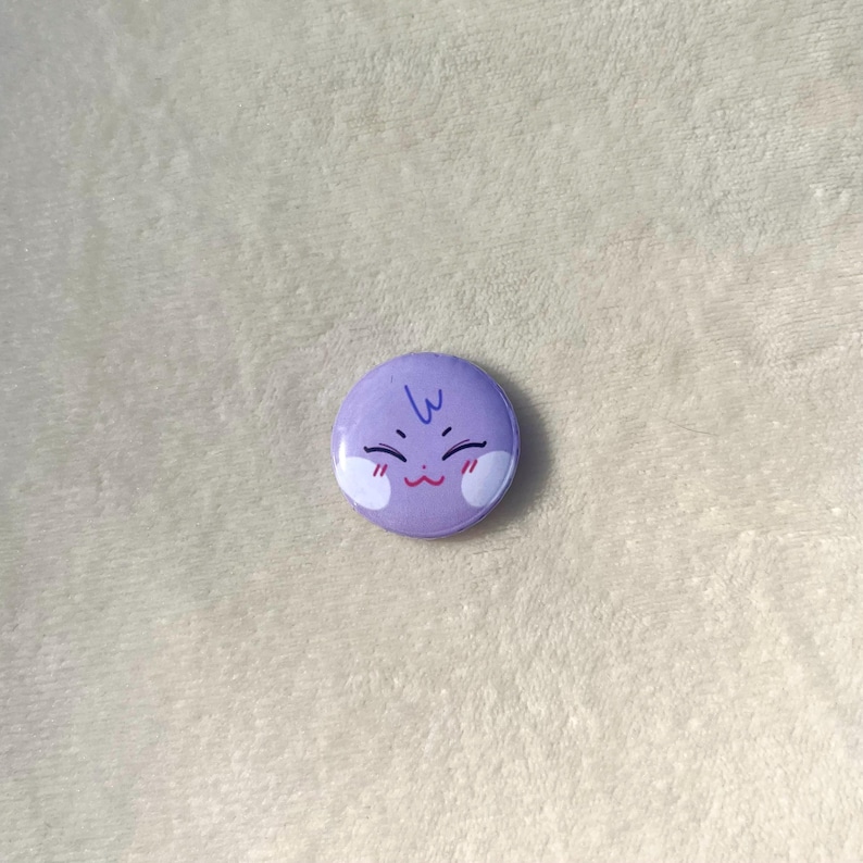 Puede incluir: Bot&oacute;n morado con una cara de dibujos animados con una peque&ntilde;a sonrisa y rubor. La cara tiene dos peque&ntilde;os c&iacute;rculos blancos para los ojos y una peque&ntilde;a l&iacute;nea azul para el cabello.
