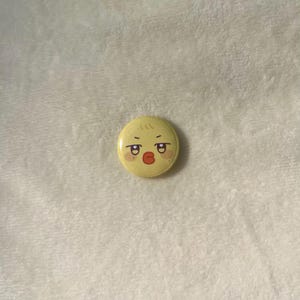 Puede incluir: Un bot&oacute;n amarillo con una cara de pollito de dibujos animados con labios rojos y rubor en las mejillas.