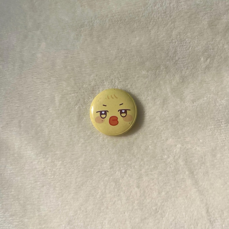 Puede incluir: Un bot&oacute;n amarillo con una cara de pollito de dibujos animados con labios rojos y rubor en las mejillas.