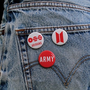 May include: Three round pinback buttons pinned to a denim pocket. One white button has red text that says "ARIRANG". Another white button has a red design. A red button has the word "ARMY" in white text.