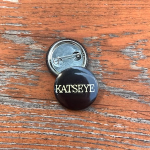 May include: A circular black pin-back button featuring the word KATSEYE printed in a bold, light-colored serif font. The back of the button is metallic silver with a horizontal pin mechanism visible, showing the construction of the accessory.